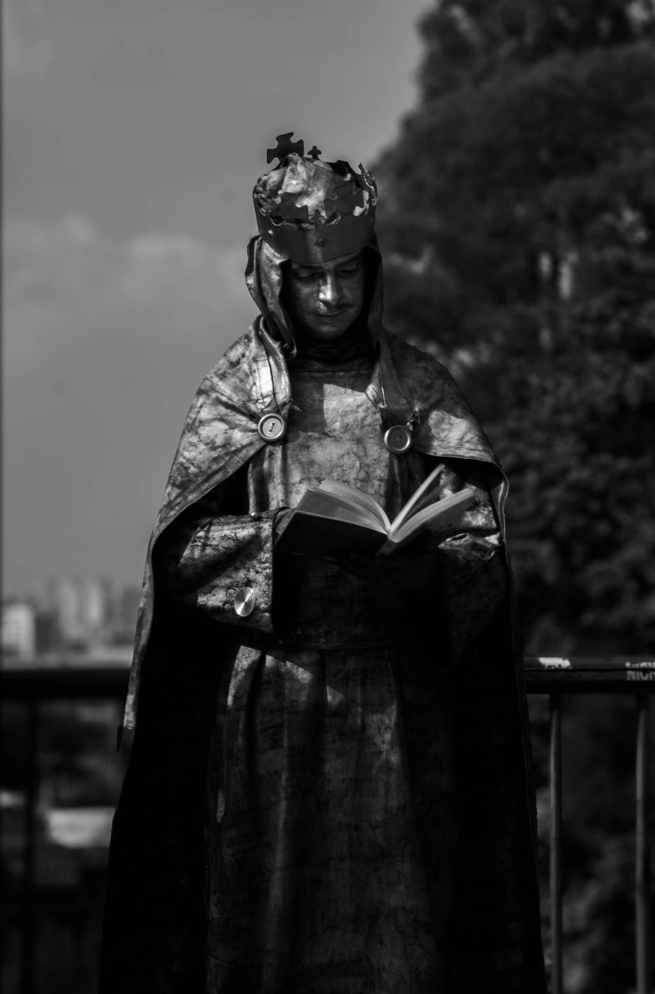 A dramatic black and white portrait of a performer dressed as a king, reading a book outdoors.